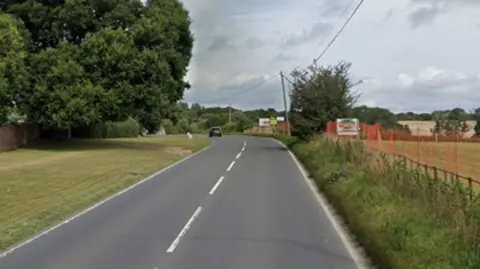 A Google Street Map image of a road. There is a large, bushy tree on the left and a field on the right.