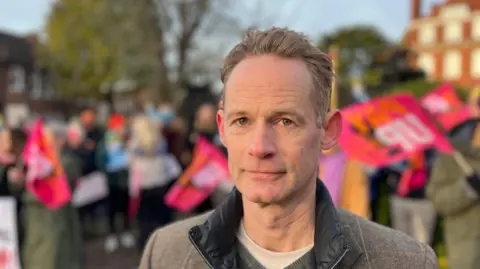 George Carden/BBC A man with short blonde hair looking at the camera. He is wearing a brown jacket. In the background people are waving red flags in front of trees and a red brick building