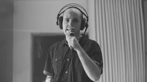 A man with a bald head, wears a headphone set and sings into the microphone in a studio 