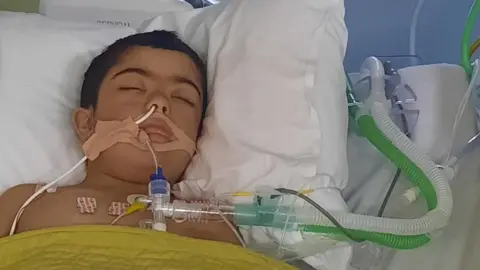 A young boy with dark hair is lying unconscious in a hospital bed with tubes coming out of his mouth and nose. A hospital monitor is at the head of his bed. A brightly coloured family blanket is on the bed cover. 