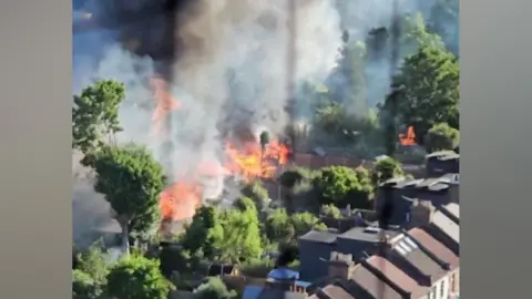 A high-angle, slightly blurred photo showing a serious fire burning intensely amongst green trees and back gardens behind a row of terraced houses, with thick columns of dark smoke filling the air.