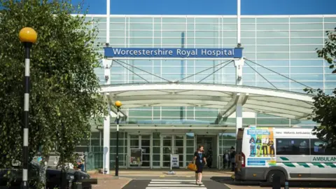 The exterior of a hospital building, with glass walls and a blue sign saying 'Worcestershire Royal Hospital'. A person crosses a zebra crossing outside the building and a silver and green ambulance is parked outside.