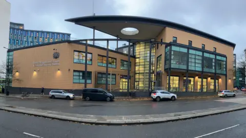 The front of Bristol Magistrates' Court, a building of three-storeys which is quite modern and built from a yellowy brick with blue-tinged windows. There are a few cars and a van waiting out the front of the building. It is a rainy day and the road looks wet.
