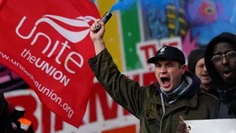 PA Media A striking bin worker wears a black cap and green coat. He is shouting and holding a blue flare in his right hand. He stands next to a red and white Unite the union flag