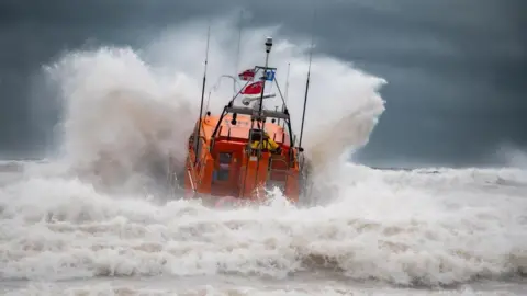 Mike Milner/RNLI An orange RNLI lifeboat, seen from behind, launches head-on into heavy seas, causing a huge white bow wave to spray up at either side of the vessel. White foaming waves crash in the foreground and a dark, stormy sky forms the background. The red ensign and flags of the RNLI hang from the boar's mast.