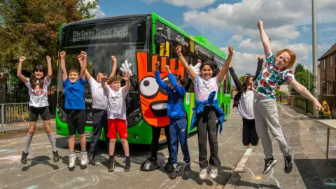 A line of children jump in the air and cheer in front of a green bus