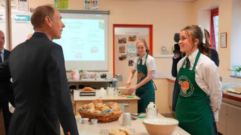 Aaron McCracken The Duke of Edinburgh, Prince Edward, in a classroom, chatting to a girl in a green apron. There are mixing bowls and loaves of bread on the bench. 