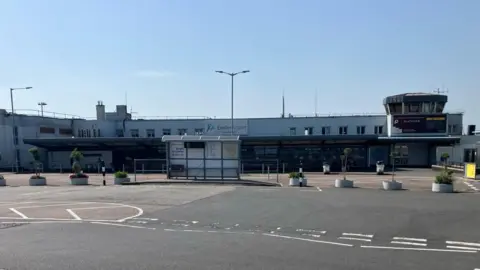 The outside of an empty looking Exeter Airport. Nobody waiting at the bus stop or any cars. There are a few people in the shade out of the direct sunlight. 