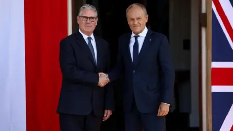 Sir Keir Starmer and Donald Tusk are stood up and shaking hands. They are both wearing dark coloured suits, white shirts and ties. They are stood in front of a Polish flag and a Union flag.
