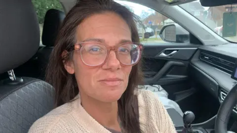 Woman with dark hair and glasses sat in a car.