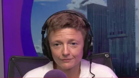 Bev Clarke sits in a studio with a purple screen behind her and a photograph of two high rise buildings. She is wearing black earphones and a white shirt and sitting in front of a purple, foam microphone cover