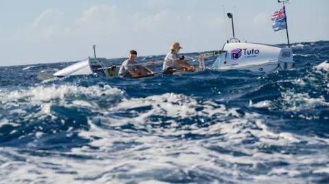 Adam Rowlatt Two men on a white rowing boat on a choppy ocean, with a logo for sponsor Tuto visible on the side. The one at the front has his face turned away from the camera and is wearing a hat and grey t-shirt. The man behind him is looking towards the camera and smiling. He has dark brown hair.