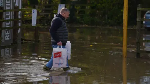 PA Media A man walking through knee-high water in a car park carrying two white plastic bags. The bag nearest the camera has "Jack's" written on it and we can see small bow waves from the front of his wellies as he moved.