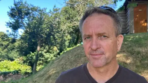 James Meller - a man in a dark grey t-shirt, and dark grey hair with blue eyes looks into the camera. Behind him is a grassy hill, trees and a brown wooden building. 