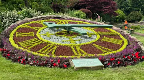 Hands and Heels A large floral clock. The hours are marked out in roman numerals with light green bedding plants, set against a background of darker ones, with light green ones making up the centre. The whole is surrounded by a border of dark purple and red flowers. Its minute and hour hand are in green metal. A plaque is set in front of the decorative structure, and on the right a park worker, in an orange florescent outfit, is dwarfed by its large scale.