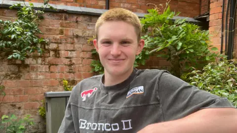 A photo of a man sitting on a bench smiling into the camera. He has got short orange bleached hair and it is very short. You can only see his head and shoulders. He has got a gray top on which has stitching on it with BRONCO written. 