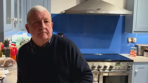 A man stands in his kitchen. He is wearing a blue jumper. The wall behind him is blue. He is leaning against a work surface.