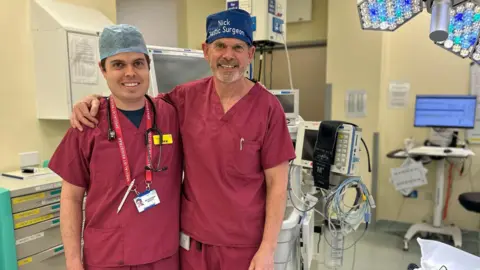 Dr Nicholas Bennett and Dr Alexander Bennett smiling for a photo at the hospital after the procedures they performed together. The father is wearing a blue had with his name on it. The son has a blue operating cap.