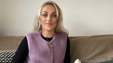 BBC Christie has shoulder-length blonde hair and is sitting on a beige sofa in a living room, wearing a lilac textured vest over a black top and patterned trousers, with decorative cushions and neutral furnishings in the background.”