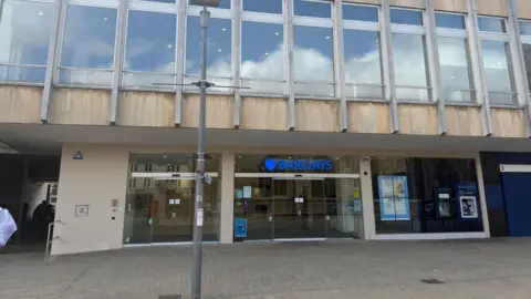The glass front of an empty Barclays bank, with a glass-fronted storey above it and a pavement in front of it.