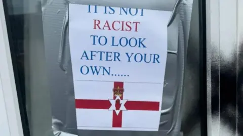 Sign taped to a window reading 'It is not racist to look after your own .....' with Northern Ireland flag below  