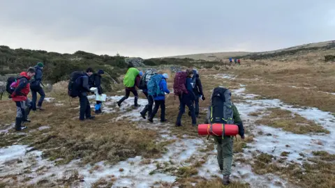 Dartmoor Plodders A group of people with rucksacks trekking over the moor which has patches of snow on it.