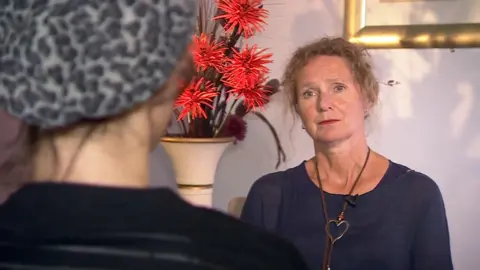 In the foreground is a young woman wearing an animal print hat and a black jumper. She is slightly blurred. In the background, in focus is a woman with light brown tied back curly hair wearing a navy blue top and a silver necklace. Next to her is a vase of orange flowers.