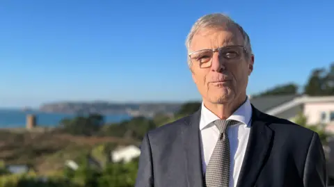 Cyril is dressed in a dark suit, white shirt, and patterned tie stands on a balcony overlooking a scenic coastal landscape. The background includes greenery, rooftops, and a view of the ocean under a bright blue sky.