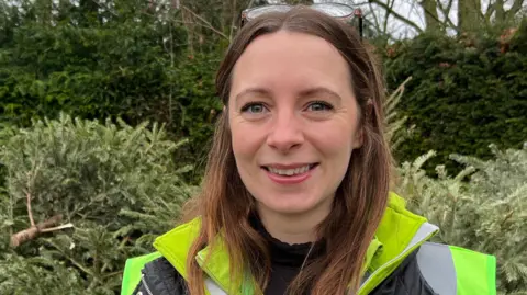 Jodie Vaughan is off centre to the right. She has long brown hair and is wearing glasses on top of her head. She is wearing a black top and a yellow hi-vis jacket. 