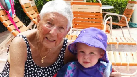 Jason Camis Ilse Camis has short white hair and tanned face and appears to be on holiday. There are sun loungers behind her. She is wearing a black swimming costume with a pattern of small white squares and is sitting with her arm around a toddler, Luciana, who is wearing a purple top and purple bucket hat.