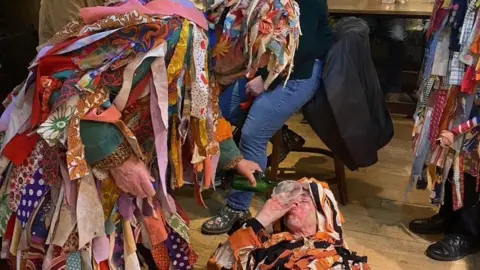 Three figures in coats covered in rags perform a play on a pub floor. One is laying on the ground with a funnel in his mouth while another pours liquid from a green bottle.