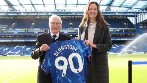 Frank Blunstone poses for a photo with Laura Cordingley, CEO of the Chelsea Foundation, whilst holding a Chelsea shirt reading "Blunstone 90" prior to the Premier League match between Chelsea FC and Newcastle United FC at Stamford Bridge on October 27, 2024 in London