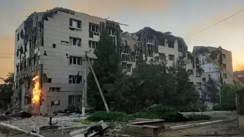 A large grey boxy building is visibly damaged and surrounded by debris and rubble. Flames are visible on one side of the building.