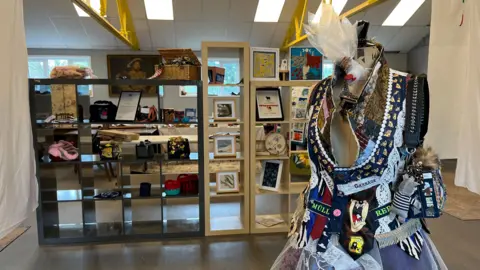 Tom Jackson/BBC A room in which there is some shelving with various items on it. In front of it is a headless mannequin with handmade jewellery and a bag hanging on it. There is also a handmade, craftsy-style waistcoat and skirt on it which has been made from various pieces of different clothing to create an entirely original piece.