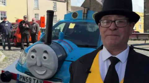 Martin Campbell, a middle-aged man dressed as the Island of Sodor's Fat Controller, complete with a black jacket, yellow waistcoat, and black top hat. He is stood next to Thomas, who has been parked in a cobbled street.