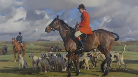 Kieron Williamson/National Horseracing Museum A painting of an English hunting meet. A male rider wears a red jacket on top of a bay coloured horse. Another rider can be seen to the left riding a chestnut horse. Several hounds sit around the horses. 