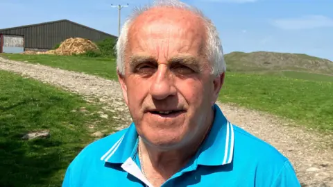 Jennie Dennett/BBC Greg Penellum, an elderly man with grey hair and thick eyebrows, stood facing the camera on his farm. In the background, there is a field of short grass that rises in the distance, as well as a dark grey farm building. Greg is wearing a bright blue polo shirt with a twin-tipped white collar, and a small chain can be seen through the opening.