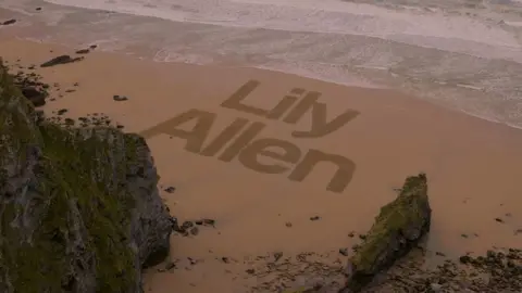 Boardmasters The name "Lily Allen" is written out in the sand of a beach as waves roll in.  