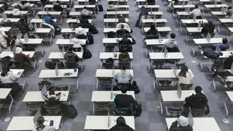 YONHAP/EPA/Shutterstock Students sat in rows on desks about to take an exam. Grey flooring.