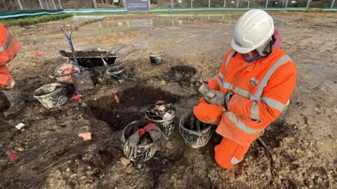 George Carden/BBC A man in a white hardhat and orange high-vis jacket and trousers on a muddy surface looking at an old glass bottle.