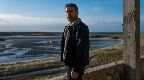 Dónall Ó Héalai  in a denim jacket and jeans, pictured in front of a Donegal bay