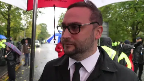 Christian Wakeford, who has light brown hair and short facial hair, and is wearing a black jacket and white shirt, speaks to the camera while holding a red and white umbrella 