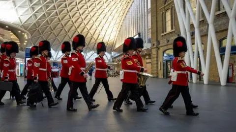 PA Media The guards marching through Kings Cross train station's concourse in full uniform. Most are holding musical instruments. The concourse is largely empty.