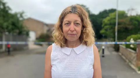 Frances Pulley a brown haired woman wearing a white top and stood in a street with a police cordon behind her.
