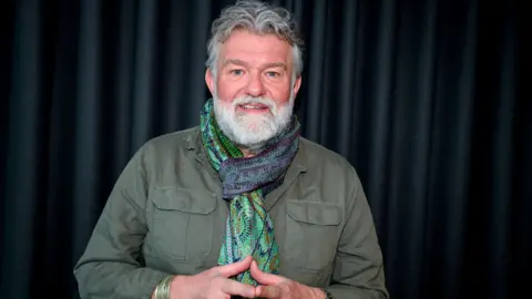 Sarah Jeynes/BBC Si King smiling into the camera in front of a dark, pleated curtain. He is wearing a green, blue and purple patterned scarf tied around his neck. He is wearing a green khaki shirt with large pockets on both sides of his chest. He is holding his hands together in front of his chest.
