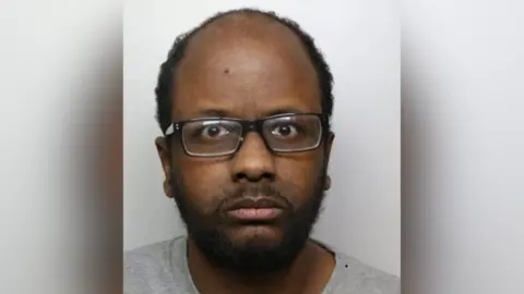 A mugshot of Zakaria Mohammed, who is wearing a grey T-shirt and black-rimmed glasses.