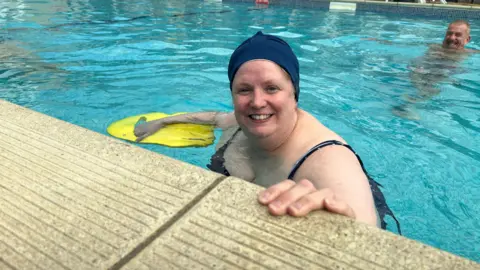 A woman standing in a swimming pool and smiling. She wears a dark swimsuit and swimming cap and holds a yellow float in her left hand.