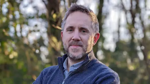 Rachel Andre Dan is stood outside with trees behind him. He has grey short hair and a beard. He is wearing a grey quarter zip jumper and a checkered shirt.