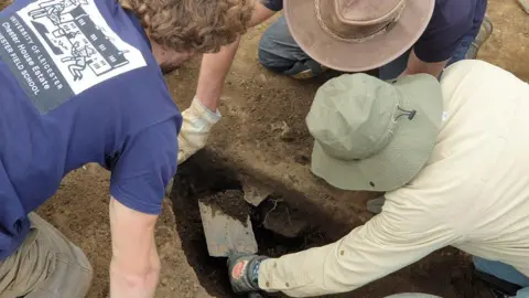 North Northamptonshire Council A figure in a beige shirt and olive hat shovels dirt out of a hole while two men in navy t-shirts crouch close by looking on.