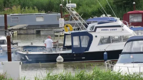 A white and blue small boat, called Sorel Light, is on a river, with greenery in the background. A man is standing on the boat with his back to the camera.